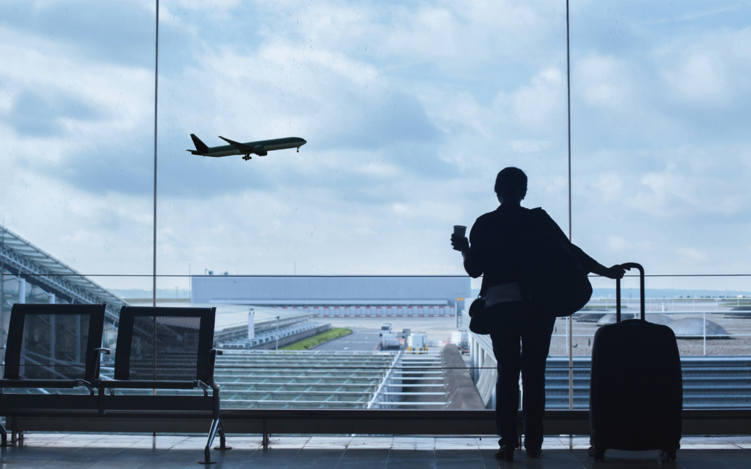 The image shows a silhouette of a traveler at an airport terminal, standing near a large window and gazing at a plane taking off or landing outside. The traveler is holding a cup in one hand and has a suitcase in the other, suggesting they are preparing for a journey or reflecting on an upcoming adventure. The scene captures the quiet anticipation of 2025 travel trends, framed by the expansive view of the tarmac and the cloudy sky.